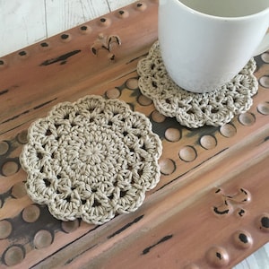 May include: Two beige, handmade crochet coasters with a floral design. One coaster is under a white mug. The coasters are made of a natural fiber and sit on a distressed, rusty brown surface.