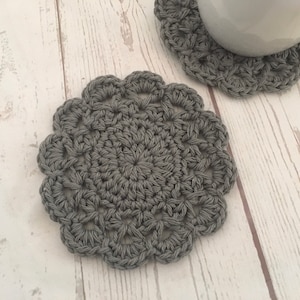 May include: Two grey crocheted coasters with a delicate floral pattern. The coasters are made of yarn and have a soft, textured surface.