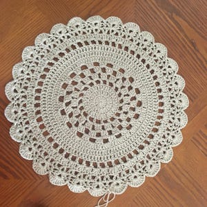 May include: A round, light beige crochet doily featuring a dense, circular centre pattern that transitions into an open, airy lattice design. The outer edge is finished with a decorative scalloped border, and a small loose yarn tail hangs below.