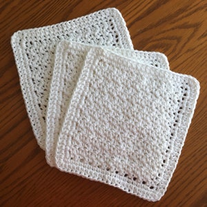 May include: Three white, square, crocheted washcloths are stacked on a wooden surface. The washcloths have a textured, handmade appearance with visible stitch patterns. The edges are neatly finished with a decorative border.