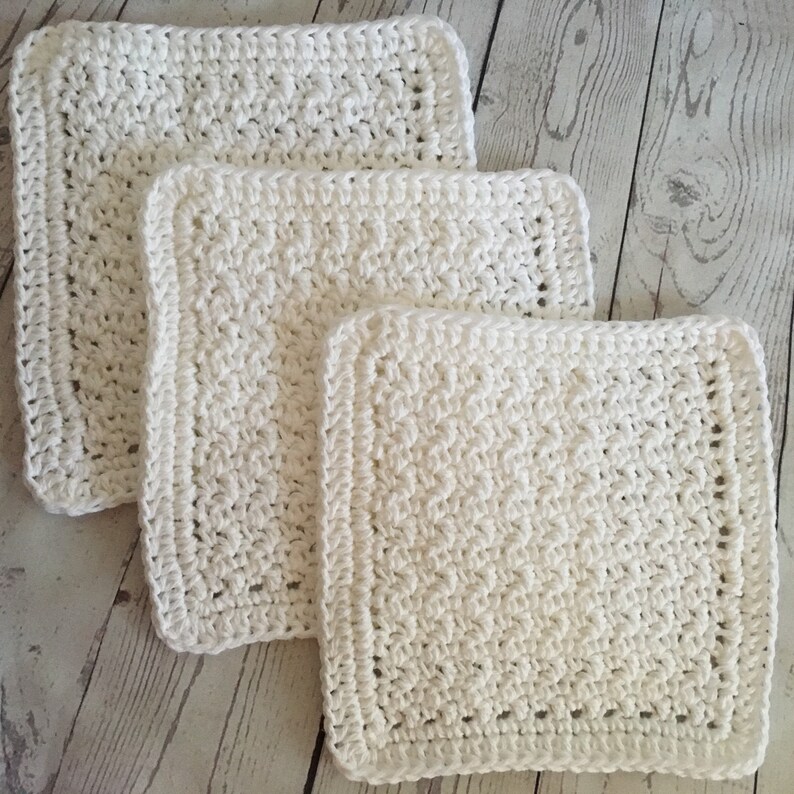 May include: Three white, square crochet washcloths are stacked on a light wood surface. The washcloths are made with a textured stitch and have a simple border. The sizes vary, with the largest on top and the smallest at the bottom.