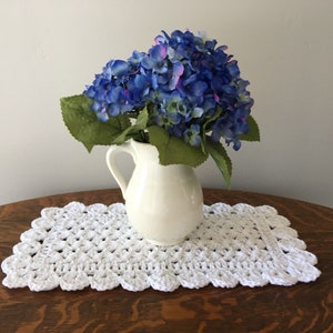 May include: A white ceramic pitcher holding blue and purple hydrangeas sits on a white crocheted table runner with a scalloped edge. The runner is placed on a dark wooden table.