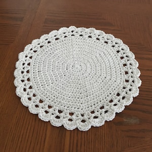 May include: White crocheted doily with a delicate lace pattern. The doily is round and has a scalloped edge.