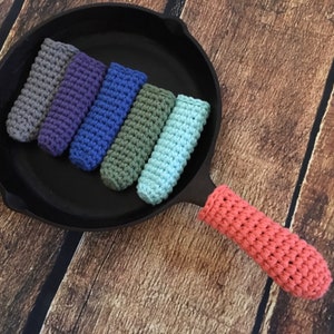 May include: A set of six colourful crocheted pot holders in various shades of blue, green, purple, and grey. The pot holders are arranged in a row on a black cast iron skillet. The skillet has a wooden handle with a crocheted cover in a coral colour.