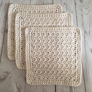May include: Three off-white, square crochet washcloths are stacked on a light wood surface. The washcloths have a textured, handmade appearance with a detailed border. The neutral color and simple design suggest a focus on functionality and natural materials.