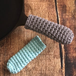 May include: Two crocheted pot holders, one gray and one light blue, are shown on a wooden surface. The pot holders are made with a simple stitch pattern.