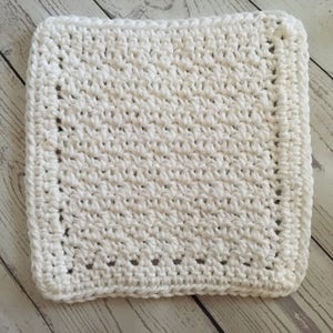 May include: A square, white crocheted washcloth. The washcloth has a textured surface created by the crochet stitches. The edges are finished with a neat border. This handmade item is suitable for use in the bathroom or kitchen.
