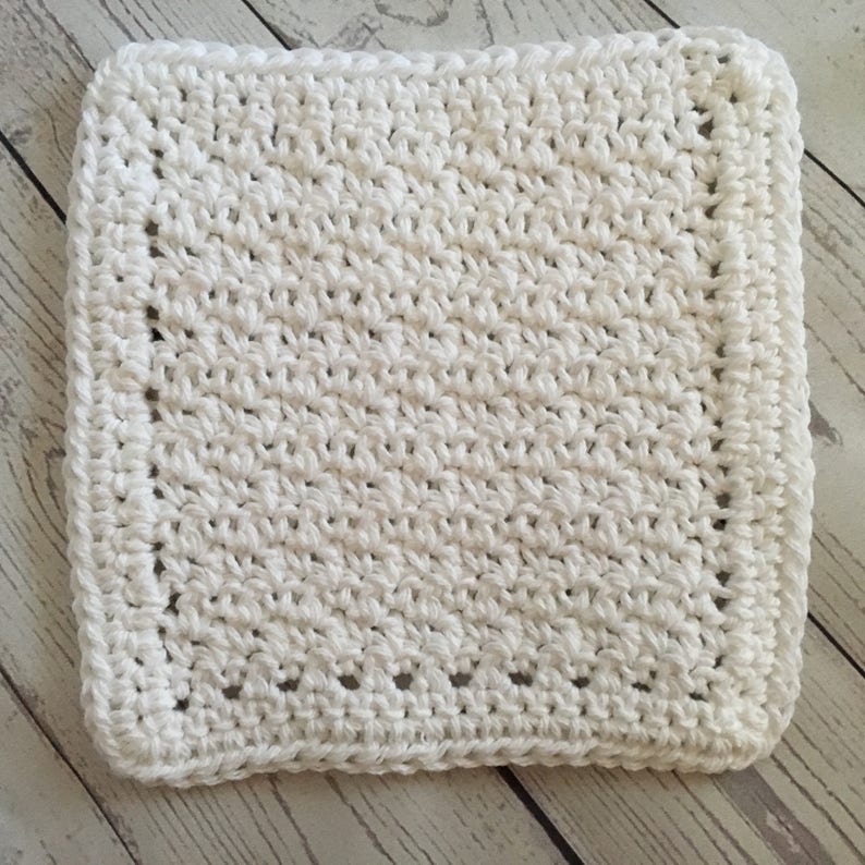 May include: A square, white crocheted washcloth. The washcloth has a textured surface created by the crochet stitches. The edges are finished with a neat border. This handmade item is suitable for use in the bathroom or kitchen.