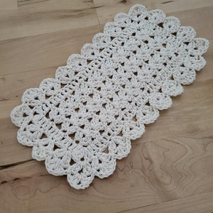 May include: A white crocheted rectangular rug with a scalloped edge. The rug is made with a simple stitch pattern and has a delicate, airy feel.
