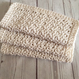 May include: Cream-colored, folded crochet washcloth. The washcloth is made of thick yarn, with a textured stitch pattern. The edges are neatly finished, and the washcloth is folded in half.