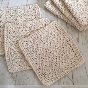 May include: Several off-white, square crochet washcloths are arranged on a light-colored surface. The washcloths have a textured appearance with visible stitches and a finished border. Some are stacked, while others are laid flat.