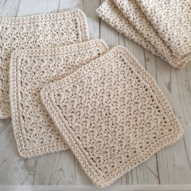 May include: Several off-white, square crochet washcloths are arranged on a light-colored surface. The washcloths have a textured appearance with visible stitches and a finished border. Some are stacked, while others are laid flat.