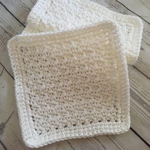 May include: Two white, square, crocheted washcloths. The washcloths are made of a textured, looped yarn and have a solid border. One washcloth is partially covering the other. The washcloths are on a light-colored wooden surface.