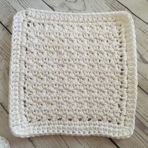 May include: A square, white crocheted washcloth. The washcloth has a textured surface created by the crochet stitches. The edges are finished with a neat border, giving it a clean and polished look. The washcloth is likely handmade.