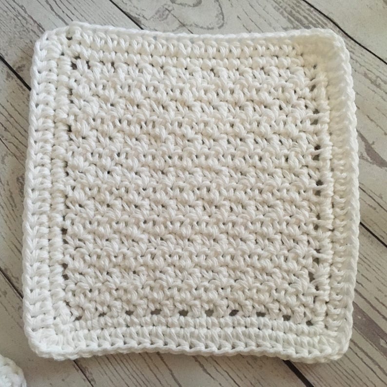 May include: A square, white crocheted washcloth. The washcloth has a textured surface created by the crochet stitches. The edges are finished with a neat border, giving it a clean and polished look. The washcloth is likely handmade.