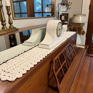 May include: A white mantel clock with a cream-colored crocheted runner on a wooden piano. The clock has a white face with black Roman numeral markings and black hands.