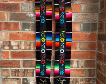 May include: A graduation stole with a black base, adorned with colorful stripes and floral embroidery in red, white, blue, and yellow. The stole has the text "Nayeli Burciaga" and "Class of 2026" embroidered on it, and is finished with orange tassels.
