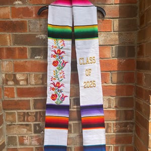 May include: White graduation stole with colorful striped patterns and floral embroidery. The stole has the text "CLASS OF 2026" embroidered in gold. The stole is hanging on a black hanger against a brick wall.