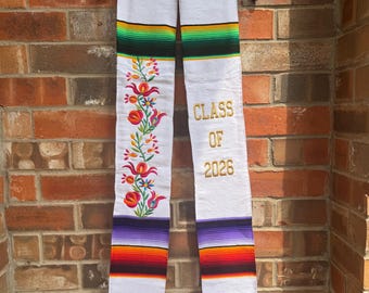 May include: White graduation stole with colorful striped patterns and floral embroidery. The stole has the text "CLASS OF 2026" embroidered in gold. The stole is hanging on a black hanger against a brick wall.