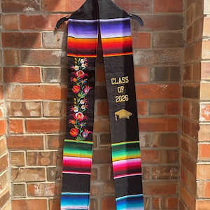 May include: A black graduation stole with colorful striped patterns and floral embroidery. The stole has the text "CLASS OF 2026" and a gold graduation cap icon. The stole is hanging against a brick wall.