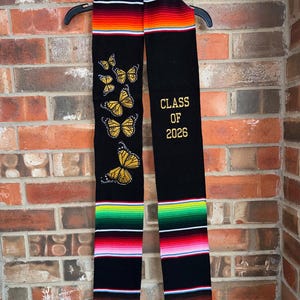 May include: A black graduation stole with gold embroidered text reading "CLASS OF 2026". The stole features a colorful striped design at the top and bottom, with embroidered yellow butterflies on one side. The stole is hanging on a black hanger.