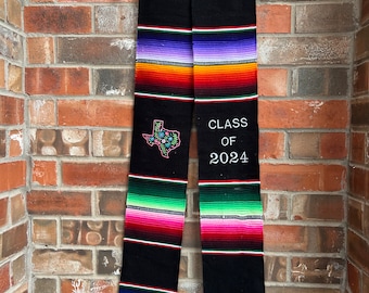 May include: Black graduation stole with colorful stripes and a white tassel. The stole has an embroidered Texas state outline with flowers and the text "CLASS OF 2024".