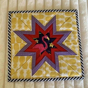 May include: Square quilted coaster with a star design. The center features a pink flamingo on a navy background. The star is red and purple with yellow pineapple print and a black and white striped border.