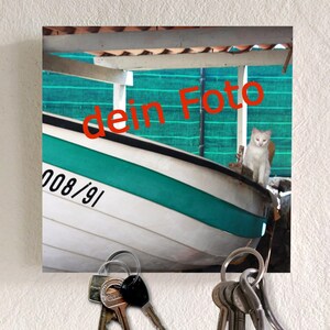 Könnte beinhalten: Ein weißes und türkisfarbenes Boot mit dem roten Text "dein Foto" ist der Mittelpunkt dieses Bildes. Eine weiße Katze sitzt auf dem Boot. Unter dem Bild befinden sich zwei Schlüsselanhänger mit mehreren Schlüsseln. Das Bild ist an einer weißen Wand befestigt.