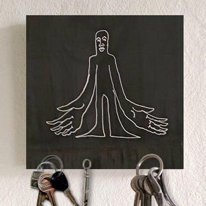 Könnte beinhalten: Ein schwarzer quadratischer Schlüsselhalter mit einer weißen Linienzeichnung einer Person mit mehreren Armen und Händen. Die Person hat ein einfaches Gesicht mit Nase und Mund. Der Schlüsselhalter ist an einer weißen Wand montiert, an der Schlüssel hängen.