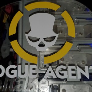 May include: A close-up of a computer case with a yellow circle and a white skull logo. The text "ROGUE AGENT" is printed in white below the logo.