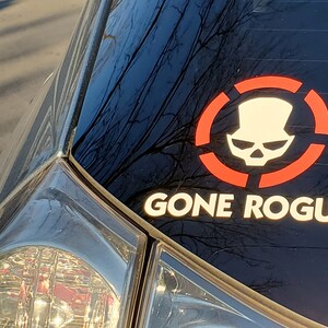 May include: A black car with a red and white sticker on the back window. The sticker features a skull wearing a top hat and the text "GONE ROGUE".