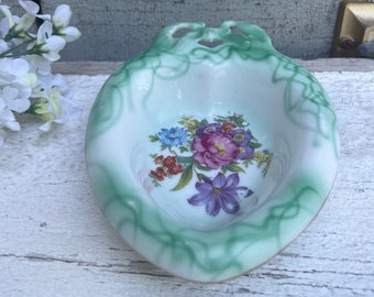 May include: A small, heart-shaped porcelain dish with a green and white floral design. The dish has a scalloped edge and a raised rim.
