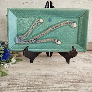 May include: A rectangular, turquoise ceramic tray with a textured surface and abstract, dark blue and tan designs. The tray is displayed on a dark brown stand against a white wooden background. Decorative floral accents are visible.