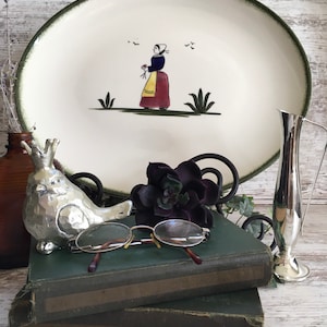 May include: A white oval serving platter with a green rim and a painted design of a woman in a red and yellow dress holding flowers. The woman is standing in a green field with green plants and birds flying overhead.
