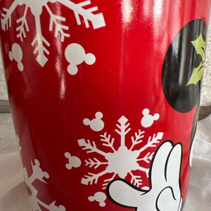 K&ouml;nnte beinhalten: Rote Keramiktasse mit einem wei&szlig;en Schneeflocken- und Mickey-Mouse-Motiv. Die Tasse hat einen wei&szlig;en Rand und einen wei&szlig;en Henkel.