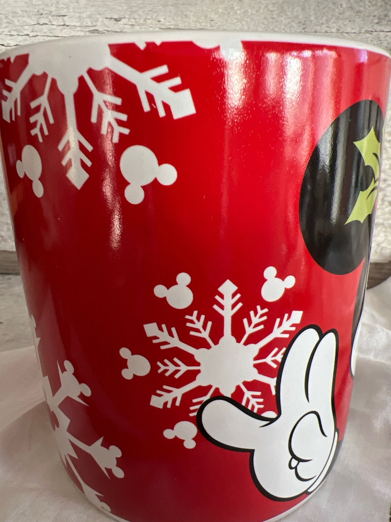 K&ouml;nnte beinhalten: Rote Keramiktasse mit einem wei&szlig;en Schneeflocken- und Mickey-Mouse-Motiv. Die Tasse hat einen wei&szlig;en Rand und einen wei&szlig;en Henkel.