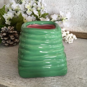 May include: A green ceramic vase with a textured, ridged exterior. The interior is a soft pink. The vase has a square shape with rounded corners. The background includes white flowers and a pinecone.