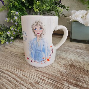 Peut inclure: Un mug en céramique blanche avec une anse en forme de cœur, orné d'une illustration imprimée d'un personnage blond vêtu d'une tenue bleu clair. Le motif comprend des arbres blancs stylisés et de petites feuilles orange et rouges à la base.