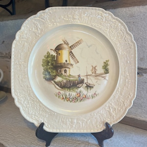 Könnte beinhalten: Ein quadratischer, cremefarbener Teller aus Keramik mit einem geprägten Blumendesign am Rand. Die Mitte des Tellers zeigt eine gemalte Szene mit einer Windmühle, einem kleinen Boot und einer Figur, die auf einem Steg steht.
