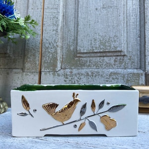 以下が含まれることがあります： 緑色の縁取りのある白い陶器製のプランター。プランターには、枝に止まっている鳥と葉の金と銀のデザインが施されています。