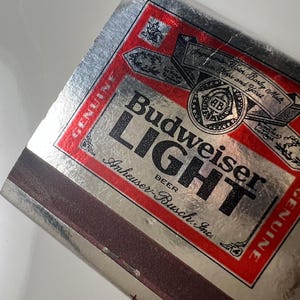 May include: A silver and red Budweiser Light matchbook. The matchbook features the Budweiser Light logo in large, bold letters. The packaging includes the words "Genuine" and "Anheuser-Busch, Inc."