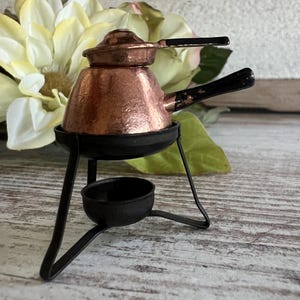 May include: A miniature copper coffee pot with a black handle and lid, sitting on a black metal stand. The pot has a hammered texture and is designed for a small stove. The background includes a white and yellow flower.