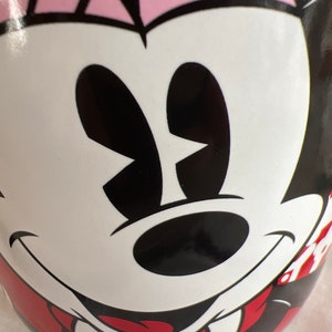 K&ouml;nnte beinhalten: Eine wei&szlig;e Keramiktasse mit einem rot-wei&szlig; gestreiften Design. Die Tasse zeigt ein Cartoon-Bild von Minnie Mouse mit schwarzen Augen, einer schwarzen Nase und einem roten Mund.