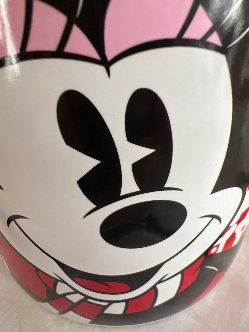 K&ouml;nnte beinhalten: Eine wei&szlig;e Keramiktasse mit einem rot-wei&szlig; gestreiften Design. Die Tasse zeigt ein Cartoon-Bild von Minnie Mouse mit schwarzen Augen, einer schwarzen Nase und einem roten Mund.