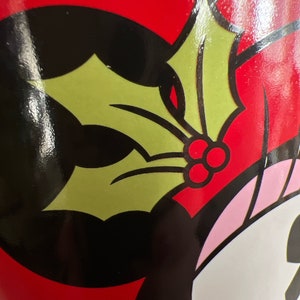 K&ouml;nnte beinhalten: Eine rote Tasse mit einer Schwarzwei&szlig;illustration einer Comicfigur, die einen gr&uuml;nen Stechpalmenzweig mit roten Beeren tr&auml;gt.