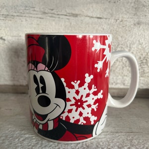 K&ouml;nnte beinhalten: Eine rote Keramiktasse mit einem wei&szlig;en Schneeflockenmuster und einem Cartoon-Bild von Minnie Mouse, die einen rosa-schwarzen Schal tr&auml;gt.