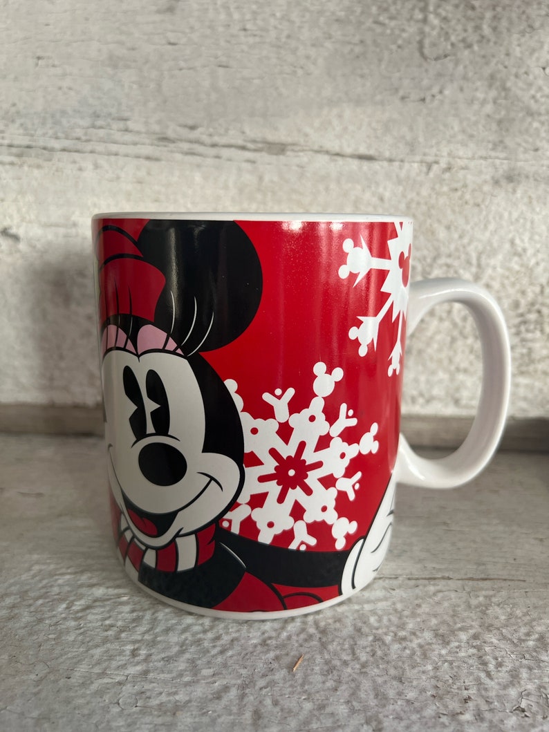 K&ouml;nnte beinhalten: Eine rote Keramiktasse mit einem wei&szlig;en Schneeflockenmuster und einem Cartoon-Bild von Minnie Mouse, die einen rosa-schwarzen Schal tr&auml;gt.