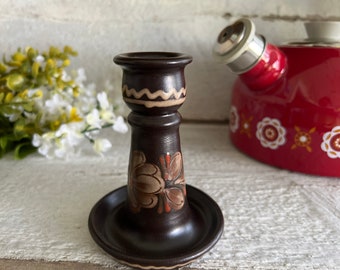 Può includere: Un portacandele in ceramica marrone scuro con un'ampia base circolare, uno stelo svasato e un bordo superiore. L'oggetto è decorato con motivi floreali dipinti a mano in tonalità terra e un motivo a linee ondulate color crema sul collo.