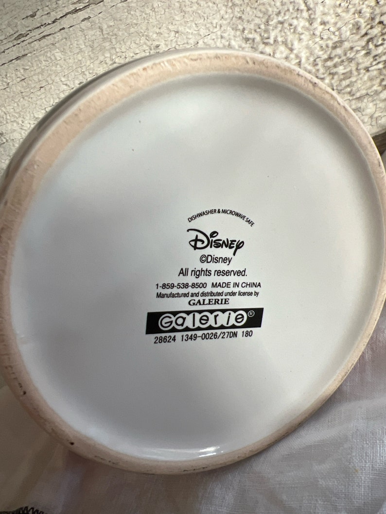 K&ouml;nnte beinhalten: Wei&szlig;e Keramiktasse mit einem Disney-Logo und dem Text "&copy;Disney Alle Rechte vorbehalten. 1-859-538-8500 HERGESTELLT IN CHINA Hergestellt und vertrieben unter Lizenz von GALERIE Galerie 28624 1349-0026/27DN 180"