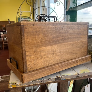 Puede incluir: Una caja rectangular de madera con acabado marrón oscuro, con un asa negra en la parte superior y asas negras en los laterales. La caja se encuentra sobre una bandeja de madera a juego. La caja se exhibe sobre una mesa rústica y desgastada.